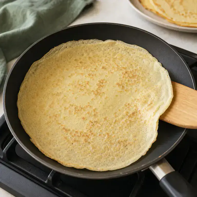 Thin golden crêpe lifted from a frying pan with a spatula